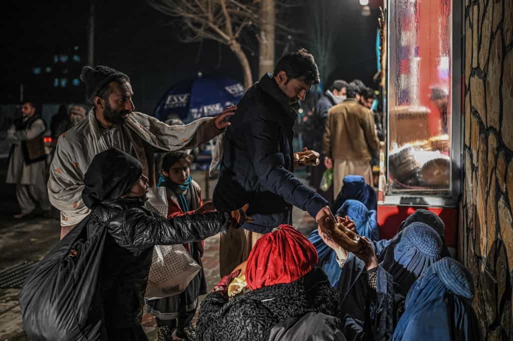 افغانستان - فقر و گرسنگی - A man distributes food to burqa clad Afghan women sitting in front of a bakery in Kabul on January 6, 2022. (Photo by Mohd RASFAN / AFP)