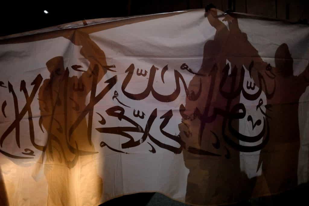 پرچم طالبان در پسزمینه، و سایه چند طالب سلاح به دست روی آن