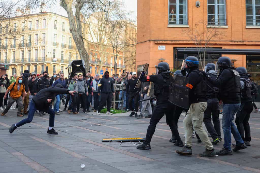 تصویر معترضان را نشان می‌دهد که به سوی مأموران امنیتی اشیاء پرتاب می‌کنند. مأموران با سپر و کلاه‌خود از خود محافظت می‌کنند.