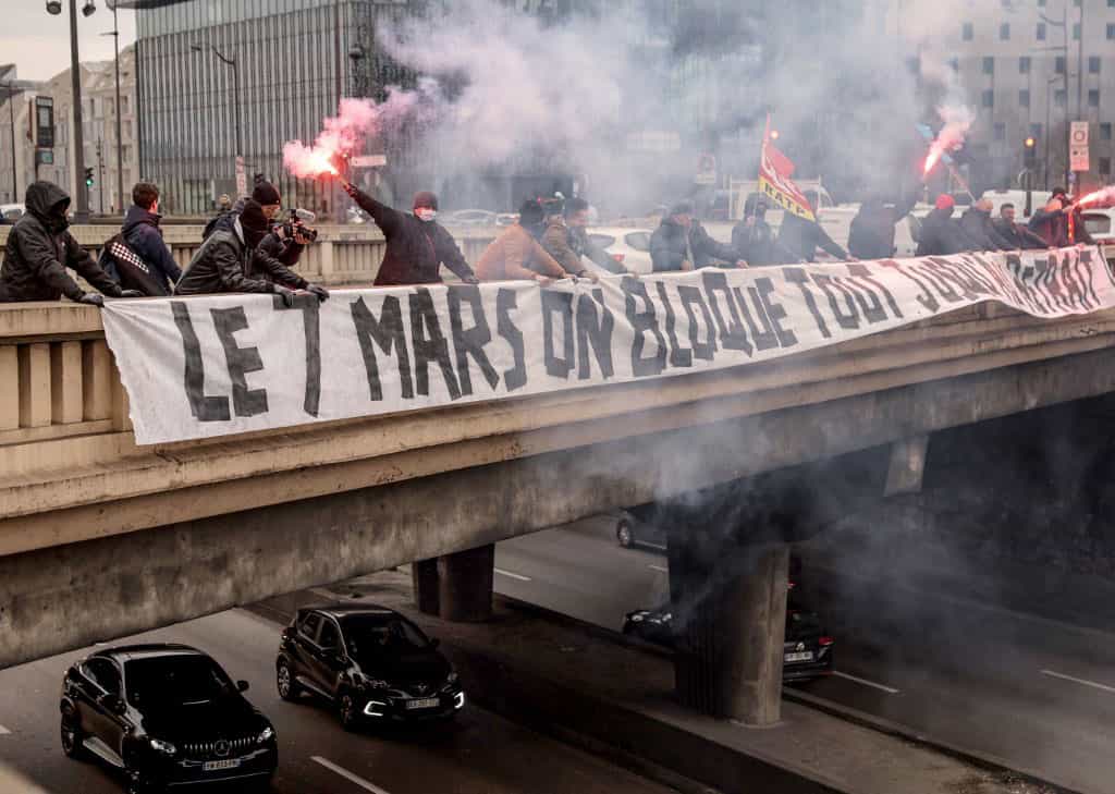 تصویر رانندگان اتوبوس شرکت دولتی RATP را نشان می‌دهد که حمل و نقل عمومی پاریس را اداره می‌کند. اعتصاب‌کنندگان در پشت بنری که به زبان فرانسوی روی آن نوشته شده «در هفتم مارس، همه چیز را تا زمان عقب‌نشینی [دولت] مسدود کنیم» بر روی پل بالای یک خیابان کمربندی پاریس قرار دارند.