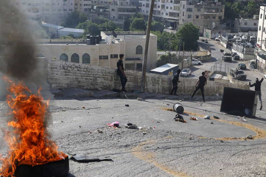 چند جوان فلسطینی در یکی از خیابان‌های سرازیری شهر نابلس در حال سنگ‌پرانی به سمت نیروهای امنیتی اسرائیل در پایین هستند. در نمای نزدیک یک لاستیک خودرو در آتش می‌سوزد.