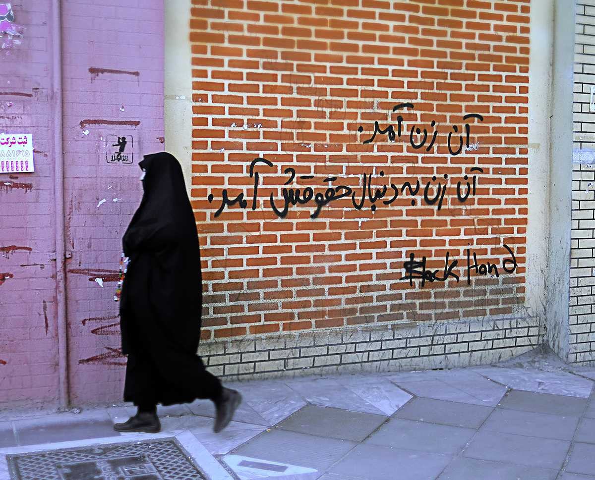 تصویری از یک گرافیتی (دیوارنگاری) با عنوان «آن زن آمد، آن زن بهدنبال حقوقش آمد». یک زن با حجاب چادر از جلوی این دیوار در حال گذشتن است.