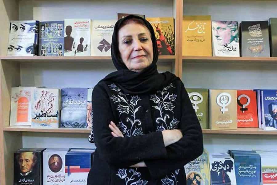 در تصویر شهلا لاهیجی مقابل یک قفسه کتاب ایستاده است.