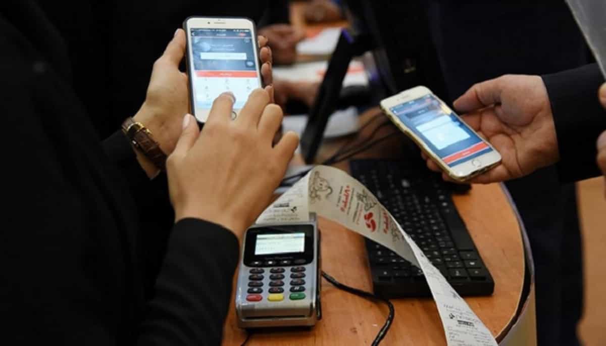 تصویری از دو نفر در حال پرداخت پول با اپلیکیشن در یک فروشگاه - روی میز یک دستگاه کارتخوان با رسید خرید دیده میشود.