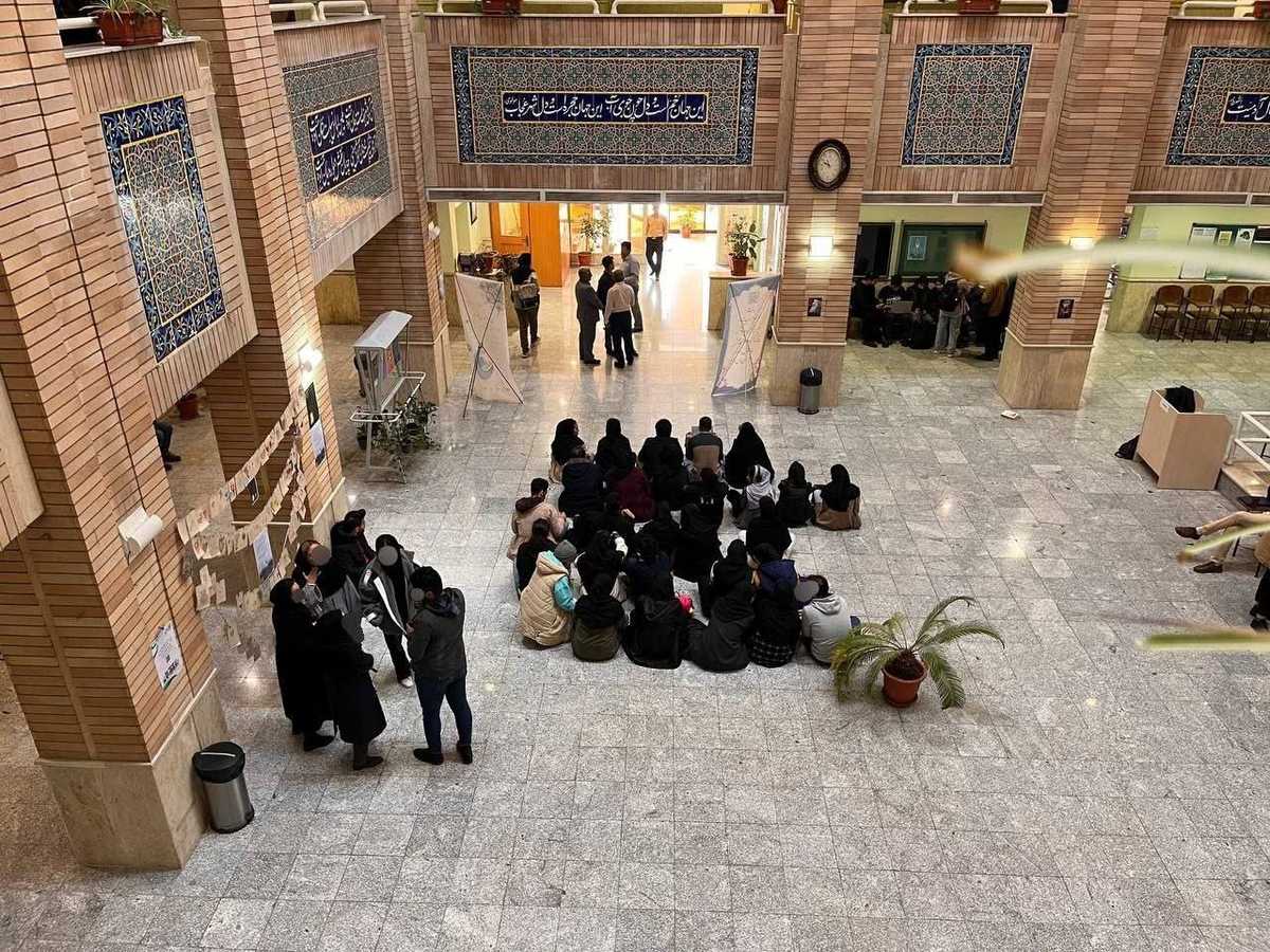 دوربین از بالا طبقه همکف دانشکده روانشناسی دانشگاه بهشتی را نشان میدهد که گروهی از دانشجویان در اعتراض به فضای امنیتی دانشگاه کف آن نشسته و تحصن کردهاند.
