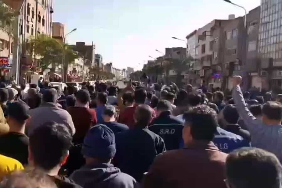 تصویر انبوه کارگران فولاد اهواز را از پشت سر نشان می‌دهند که در حال راهپیمایی‌اند. در گوشه پایین سمت راست تصویر یک کارگر مشتش را بلند کرده است.