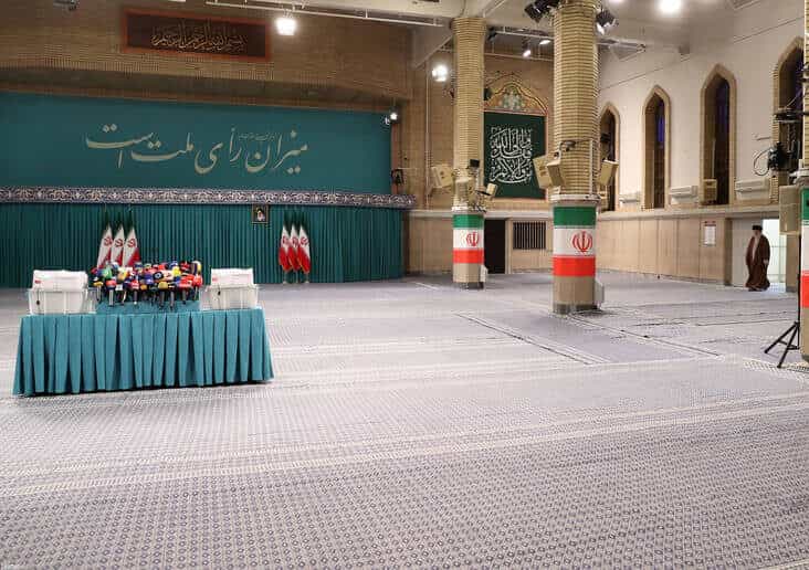 تصویر صحن حسینیه امام خمینی را نشان میدهد. در دورترین نقطه سمت راست، علی خامنهای بسیار ریز نشان داده شده که دارد از دری وارد صحن میشود. در سمت چپ تصویر، میزی که روی آن گل و