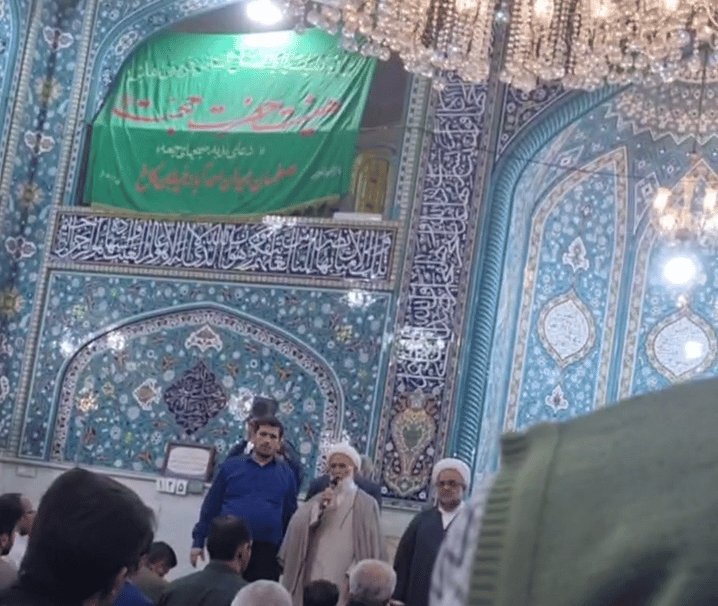 جلوی محراب مسجد حجت اصفهان دو روحانی و یک مرد با لباس آبی ایستادهاند. روحانی میکروفون به دست دارد و سخنرانی میکند