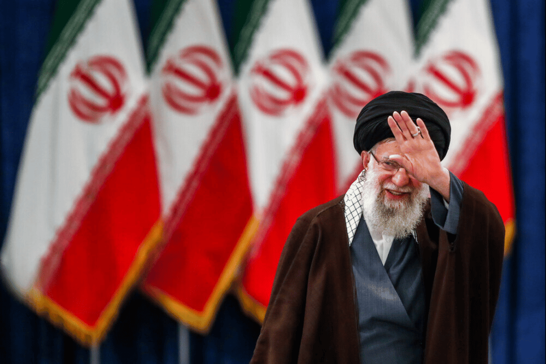خامنه ای در حال دست تکان دادن و در پشت سر او سری پرچم جمهوری اسلامی ایران