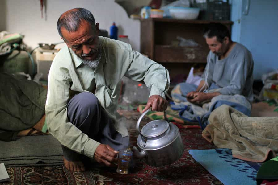 یک مهاجر سالمند افغانستانی در حال ریختن چای است و دیگری پشت سر او روی پتو نشسته و به موبایل خود نگاه می‌کند.