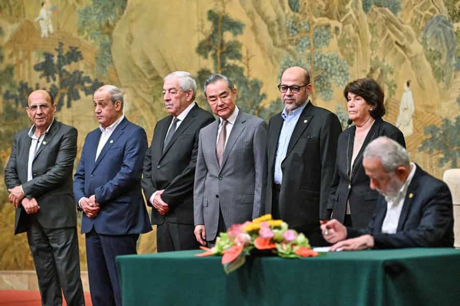 یکی از نمایندگان گروههای فلسطینی پست میز در حال امضای توافقنامه آشتی است و شش نفر دیگر در کنار او ایستادهاند. وانگ یی، وزیر امور خارجه چین در میان این جمع به میز نگاه میکند.