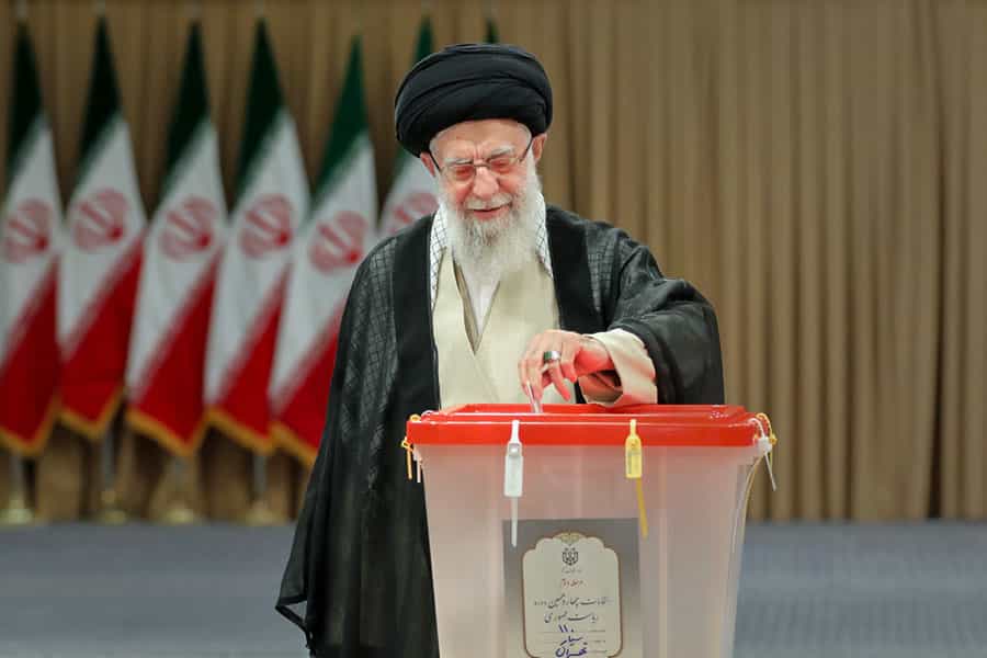علی خامنهای، رهبر جمهوری اسلامی برگه رأی را در صندوق رأیگیری میاندازد. پشت سر او چندین پرچم جمهوری اسلامی دیده میشود.