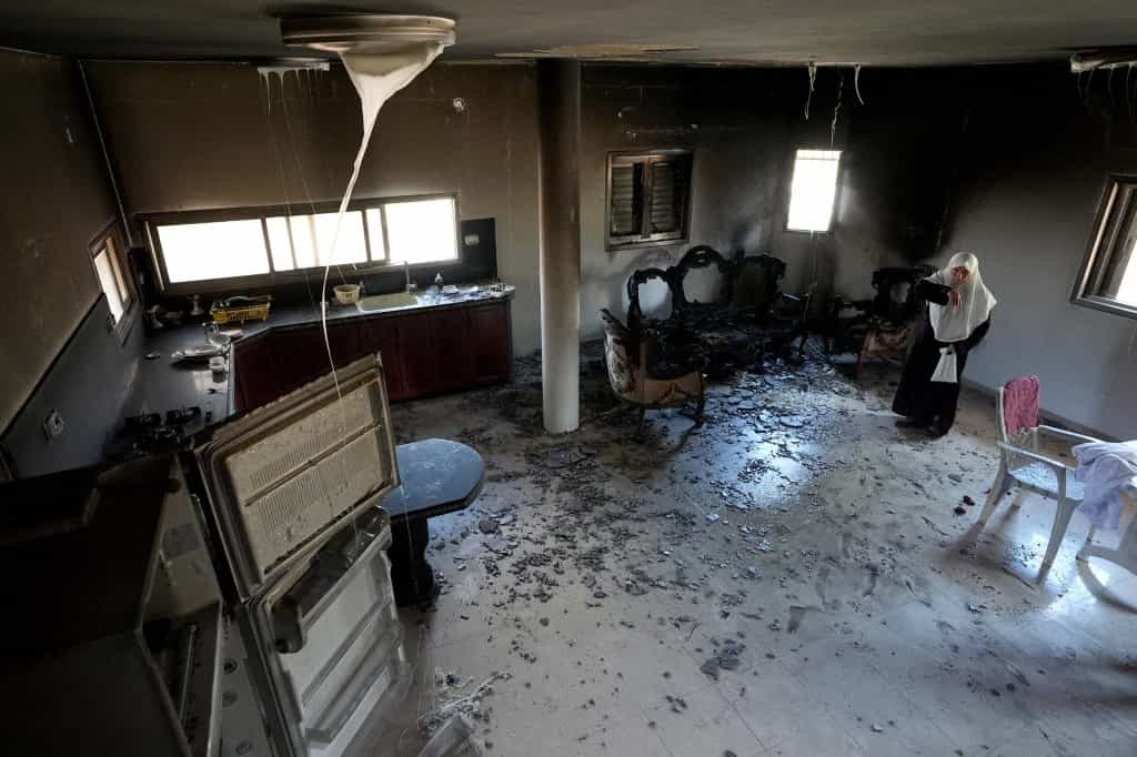 تصویر یک خانه به آتش کشیدهشده. در گوشه راست تصویر، زنی با بُهت دارد به بقایای خانه نگاه میکند.