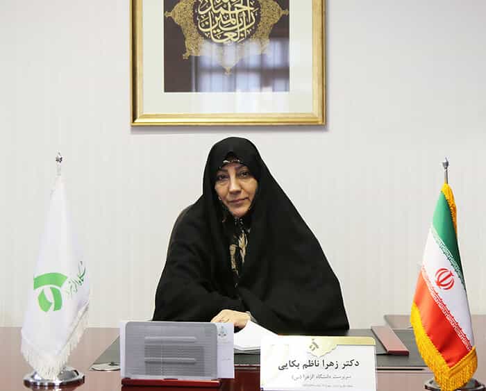 یک زن با پوشش چادر پشت یک میز مدیریت نشسته و به دوربین نگاه میکند