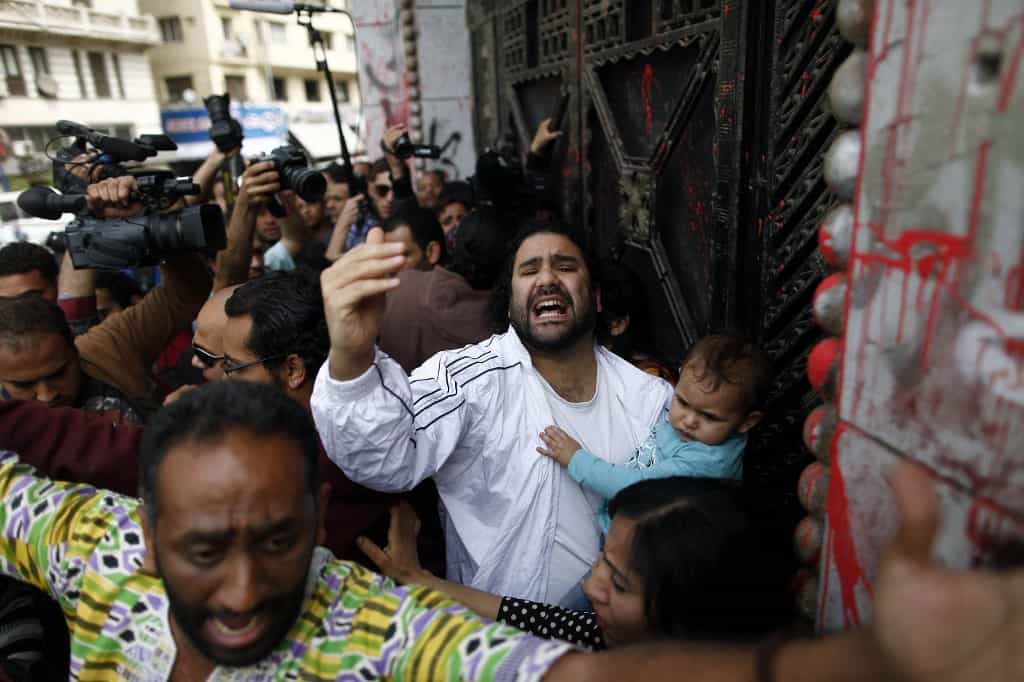 تصویر در مقابل دادگاهی در مصر در سال ۲۰۱۳ که علا عبدالفتاح فعال دموکراسی خواه مصری با فرزند کوچکش در بغل دیده میشود