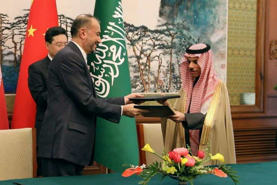 حسین امیرعبداللهیان و امیر فیصل بن فرحان، وزیران امور خارجه ایران و عربستان سعودی با لبخند بیانیه مشترکی را که امضا کردهاند با هم مبادله میکنند. پشت سر آنها پرچمهای جمهوری خلق چین، عربستان سعودی و ایران قرار دارد.
