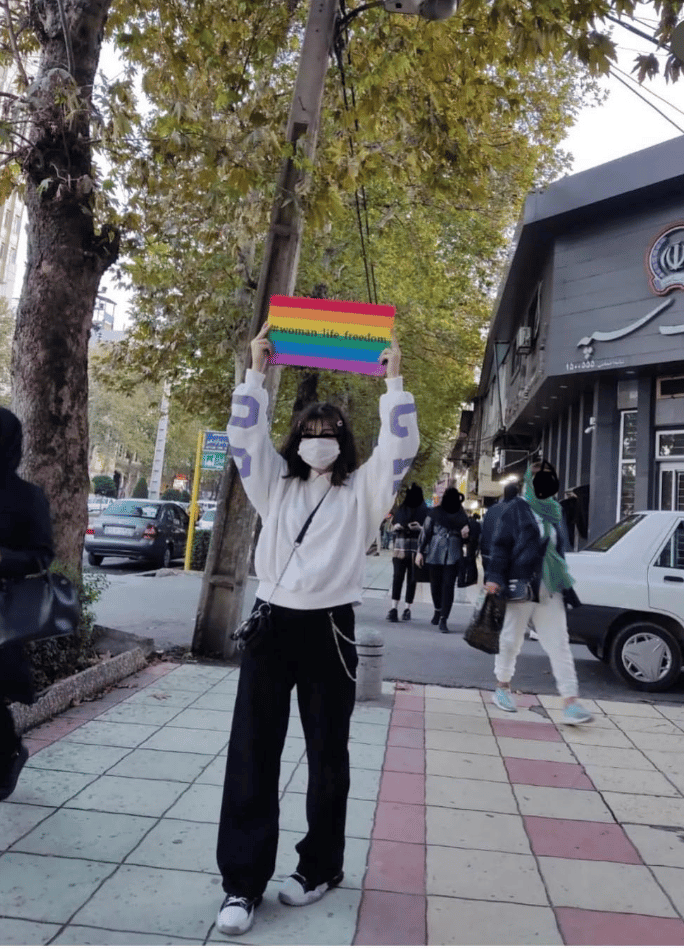 تصویری از یک فرد معترض با پرچم رنگینکمانی در یک خیابان ایران در گذر جنبش زن، زندگی، آزادی