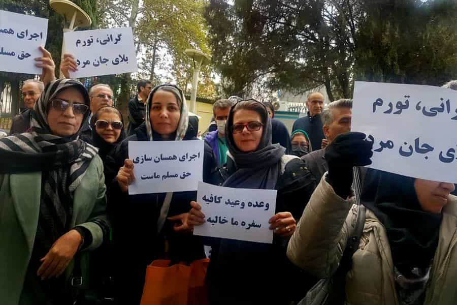 در تصویر چند نفر از بازنشستگان زن و مرد نشان داده میشوند که نوشتههایی از جمله «گرانی، تورم، بلای جان مردم»، «وعده وعید کافیه، سفره ما خالیه» و «اجرای همسانسازی حق مسلم ماست» در دست دارند.
