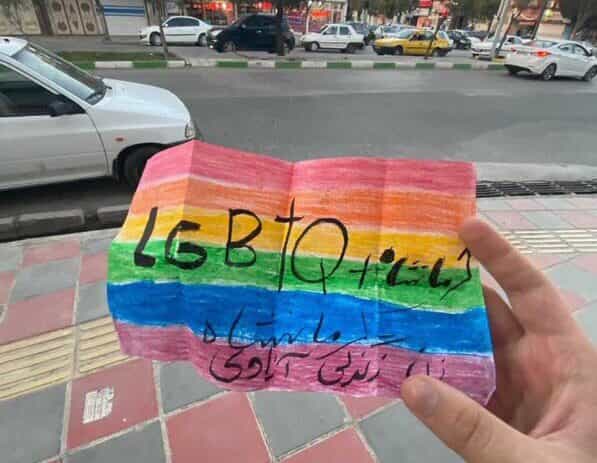 عکس تزئینی از فضای مجازی، یک دست را نشان میدهد که در یک خیابان داخل ایران، کاغذی رو به دوربین گرفته که بر آن پرچم رنگینکمان کشیده شده و نوشته شده الجیبیتیکیو پلاس کرمانشاه