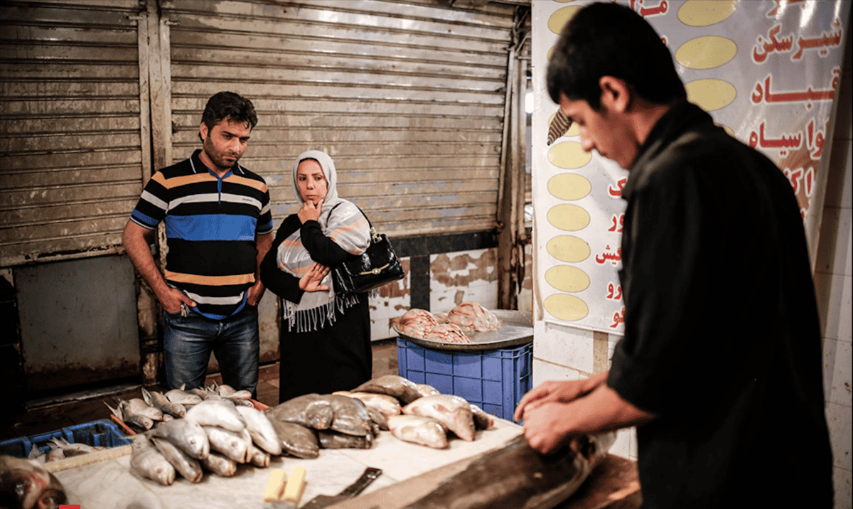 بازار ماهیفروشها - پسر جوان ماهی فروش در حال پاککردن ماهیست. یک زوج زن و مرد به ماهیها نگاه میکنند.