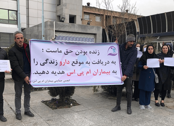 در جریان تجمع اعتراضی بیماران امپیاس، دو مرد یک بنر را دست گرفتهاند که روی آن نوشته: زندهبودن حق ماست. به دریافت به موقع دارو زندگی را به بیماران امپیاس هدیه دهید.