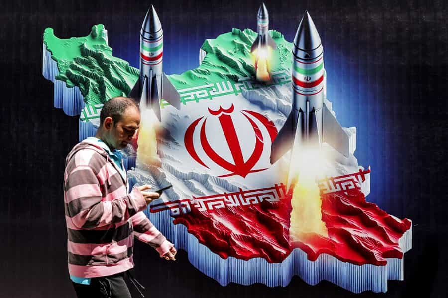 مردی در یکی از خیابانهای تهران در حالیکه با تلفن دستی خود مشغول است از کنار بنری میگذرد که در آن نقشه ایران با پرچم جمهوری اسلامی و سه موشک روی آن دیده میشود.