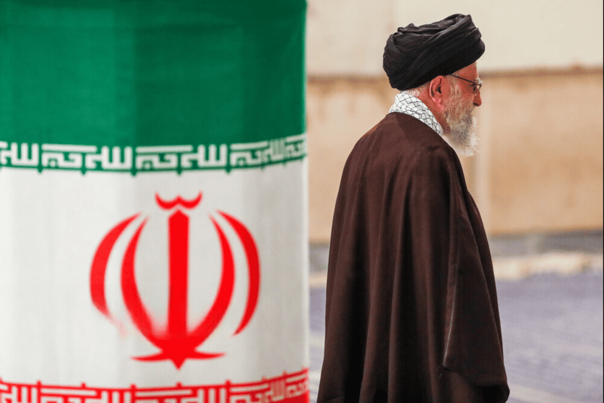 خامنه‌ای از سمت راست تصویر در حال خارج شدن است. در سمت چپ پرچم جمهوری اسلامی ایران دور یک ستون و پشت سر او دیده می‌شود