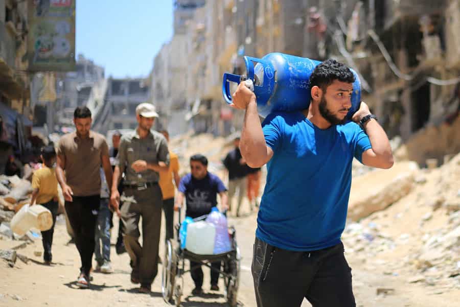 یک مرد فلسطینی کپسول گاز آبی رنگی را روی دستهای خود بلند کرده و حمل میکند. عدهای نیز پشت سر او دیده میشوند. ساختمانهای ویرانشده نیز در پسزمینه دیده میشوند.