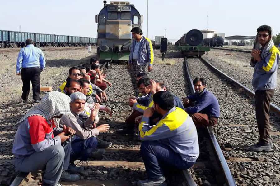 چند کارگر نگهداری خطوط راهآهن زاهدان در گرمای تابستان روی ریل قطار نشستهاند و لوکوموتیوی پشت سر آنها ایستاده است.