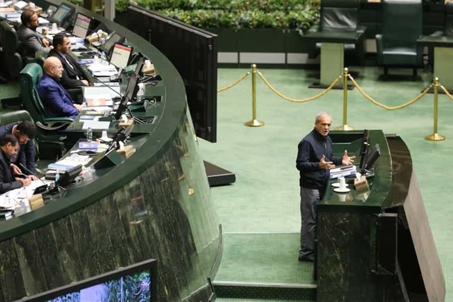 مسعود پزشکیان، رئیس جمهوری ایران پشت تریبون مجلس در حال دفاع از وزیران پیشنهادی خود است. بالای سر او اعضای هیئت رئیسه مجلس دوازدهم، از جمله محمدباقر قالیباف، رئیس مجلس دیده می‌شوند.