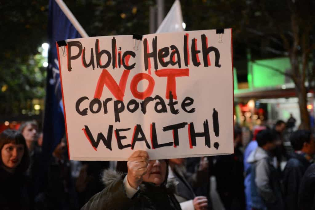 تجمعی اعتراضی در استرالیا یک نفر یک تابلو بالا گرفته که روی آن به انگلیسی نوشته: Public Health NOT Corporate Wealth (سلامت عمومی، نه ثروت شرکت‌ها)