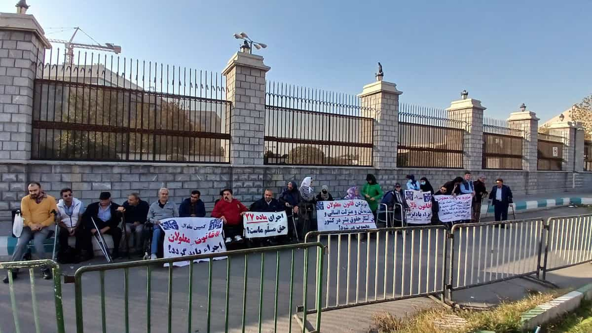 دیوار و حصارهای مجلس شورای اسلامی و افرادی جلوی آن نشسته‌اند و بنرهای اعتراضی دست گرفته‌اند