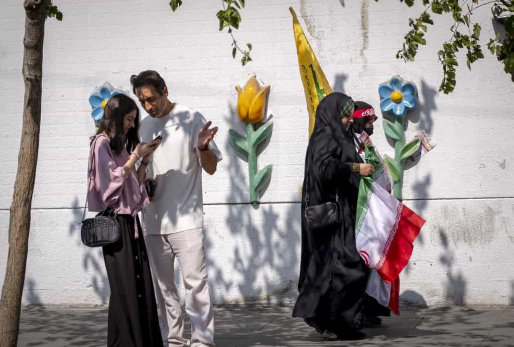 دو زن جوان محجبه که پرچم ایران و پرچم حزبالله لبنان را در دست دارند، در مرکز شهر تهران، ایران، در تاریخ ۲۷ سپتامبر ۲۰۲۴، در حال عبور از کنار دو جوان دیگر هستند که یکی از آنها حجاب اجباری ندارد، پیش از آغاز تظاهرات ضد اسرائیلی برای محکومیت حمله نظامی اسرائیل به لبنان. (عکس از مرتضی نیکوبذل / نورفوتو / از طریق خبرگزاری فرانسه)