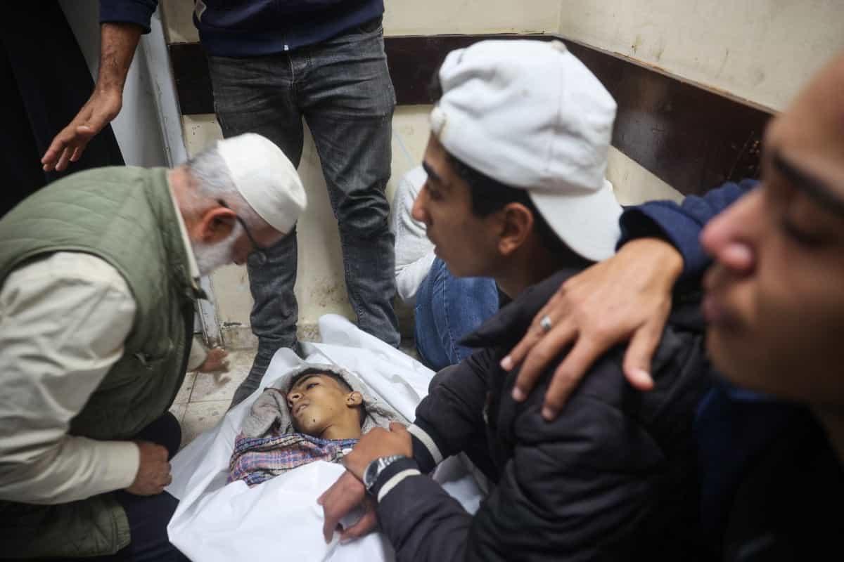 فلسطینیها بر سر پیکر کشتهشدگان حملات اسرائیل سوگواری میکنند. در وسط تصویر نوجوانی سبزهرو در میان کفن سپید پوشیده شده، و دورش را یک نوجوان دیگر با کلاه کپ سفیدرنگ که برعکس به سر گذاشته و یک پیرمرد با کلاه نخی سفید قرار دارند.