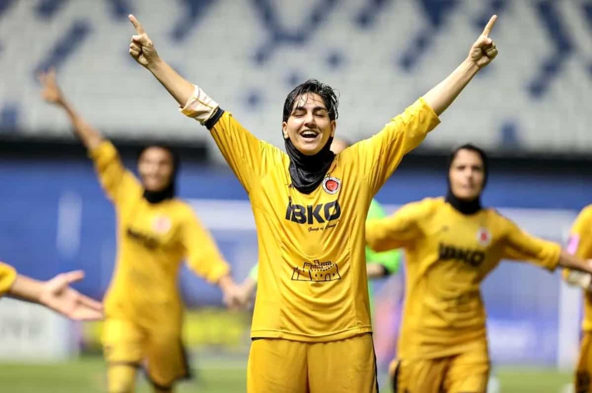 سه زن فوتبالیست با لباس زرد در زمین فوتبال شادی میکند. یکی از آنها مقنعهاش افتاده است.