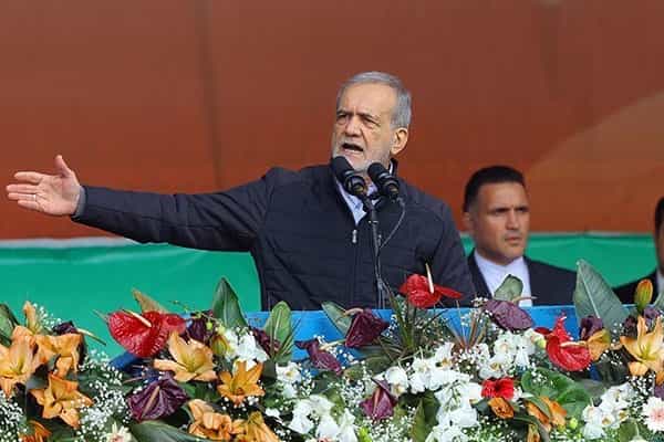 مسعود پزشکیان پشت میکروفون دست خود را بلند کرده و دارد حرف میزند. جلوی او دستههای گل دیده میشود.