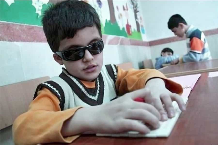 یک کودک نابینا با انگشت‌های خود کتاب درسی با خطر بریل را می‌خواند. او عینک سیاهی بر چشم دارد.