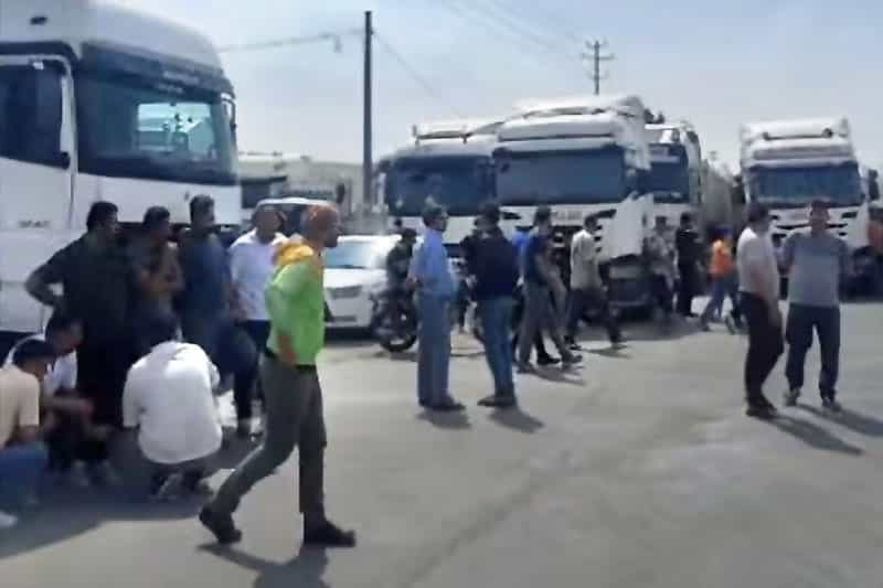 عدهای از رانندگان کامیون در هوای آفتابی در کنار کامیونهای خود ایستاده و با هم حرف میزنند.