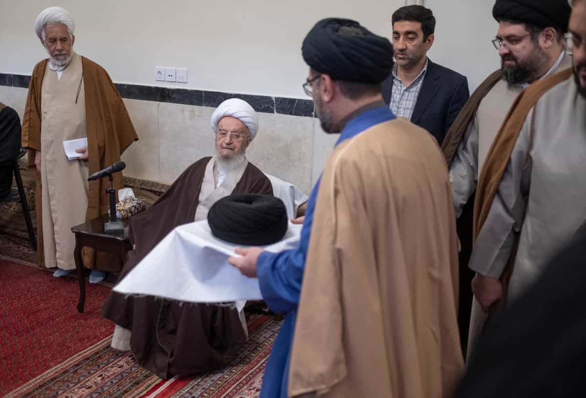 ناصر مکارم شیرازی (نفر دوم از سمت چپ) در مراسم عمامهگذاری در حوزهای علمیه در شهر قم، ۱۴۵ کیلومتری (۹۰ مایلی) جنوب تهران، در حال تماشای عمامهای است که توسط یک روحانی حمل میشود.