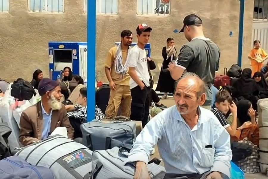 مهاجران افغانستانی در کنار وسایل زندگی خود به انتظار رسیدن اتوبوس نشسته‌اند.