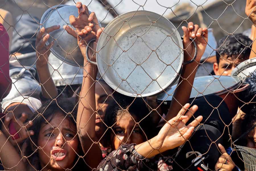 چند کودک فلسطینی پشت حصار سیمی، قابلمهها و کاسههایی در دست دارند و با نگاهی نگران منتظر دریافت غذا هستند.