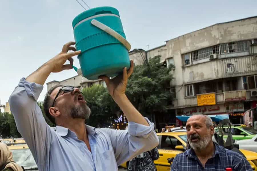 مردی در گرمای خیابان، ظرف آبی را در دست گرفته و از آن مینوشد. مرد دیگری نظارهگر اوست.