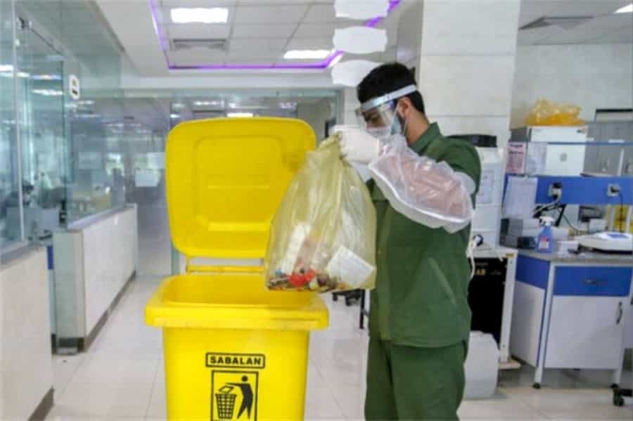 یک نفر در حال خالی کردن پلاستیک زبالههای بیمارستانی در ظرف مخصوص آن است و ماسکی بر چهره دارد.