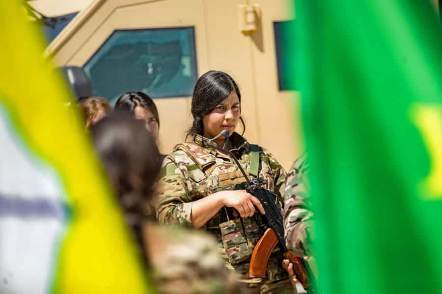 یکی از زنان مسلح «نیروهای دموکراتیک سوریه» در کنار دیگر زنان مسلح و یک خودروی نظامی ایستاده است.