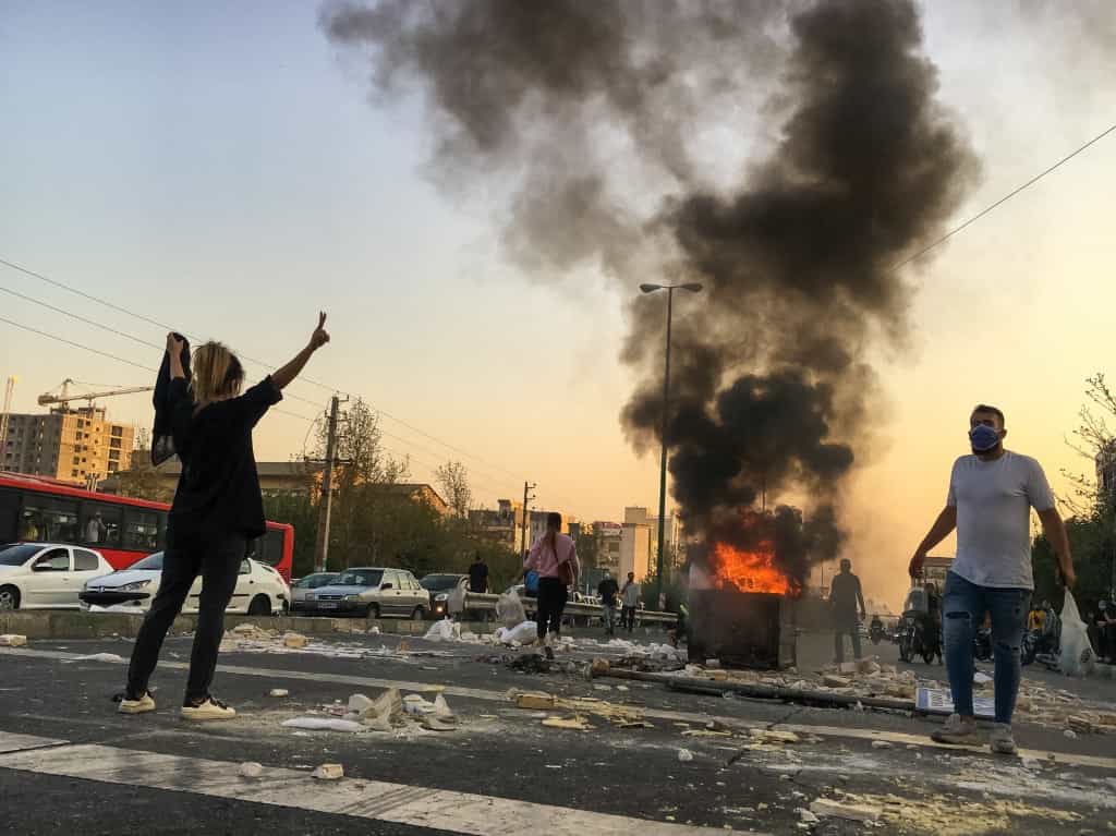 یک زن ایرانی بدون حجاب در حالی که به همراه دیگر معترضان در نزدیکی میدان پونک دور یک سطل زباله در حال سوختن جمع شده است، علامت پیروزی را نشان می‌دهد.
