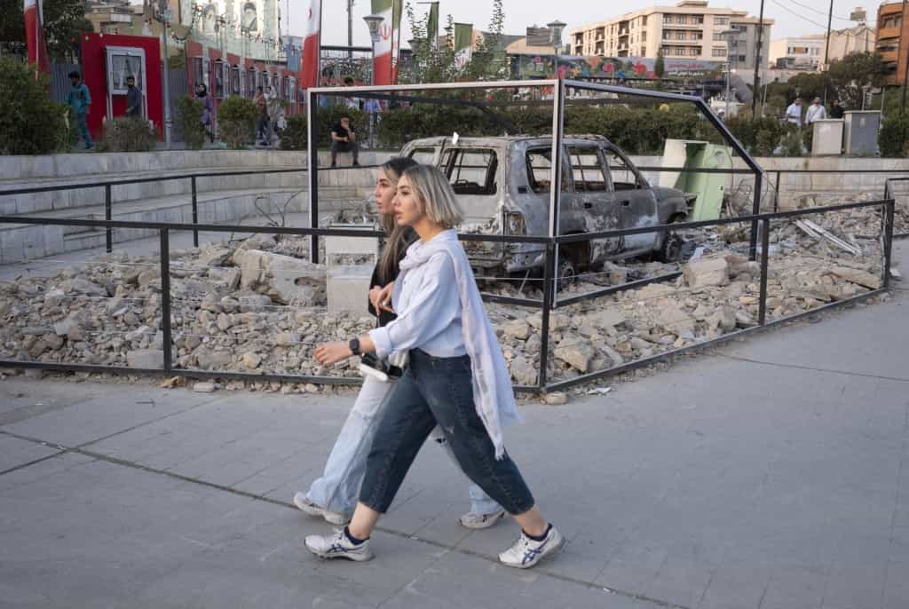 دو زن جوان ایرانی در حال عبور از کنار یک آمبولانس منفجر شده هستند. این آمبولانس بخشی از یک چیدمان ضد جنگ در یک میدان است. این اتفاق در جریان یک راهپیمایی مذهبی به مناسبت سالگرد تولد پیامبر در مرکز شهر تهران، ایران، در تاریخ ۱۹ شهریور ۱۴۰۴ رخ می‌دهد.