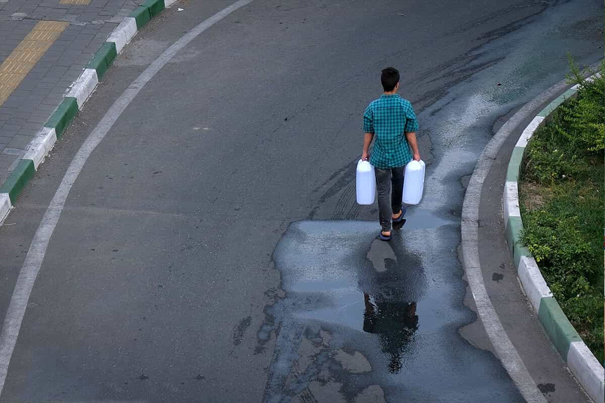 یک نوجوان با پیراهن سبز دو گالن آبردر دست پشت به دوربین راه میرود.