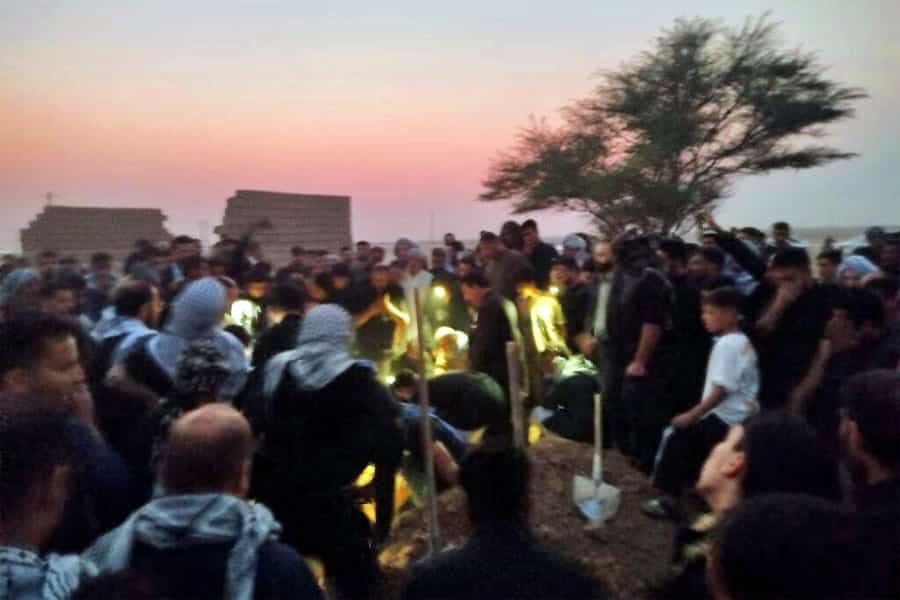 مردم در هوای نیمه‌تاریک شامگاهی در زیر نور چراغ دستی در حال خاکسپاری پیکر احمد بالدی هستند.