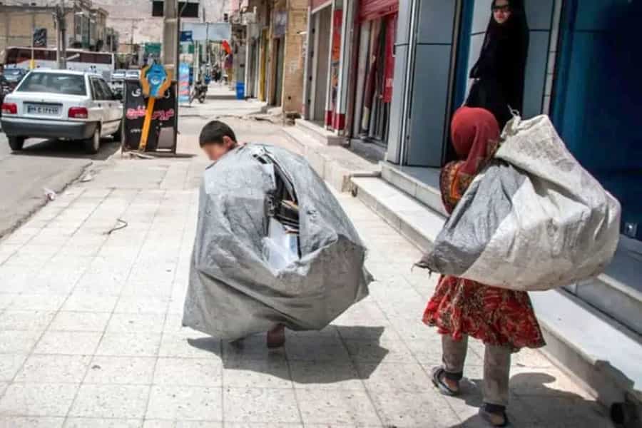 دو کودک زالهگرد در حالیکه دو کیسه بزرگ را بر دوش میکشند در پیادهروی خیابانی راه میروند. یک زن ایستاده و به آنها نگاه میکند.