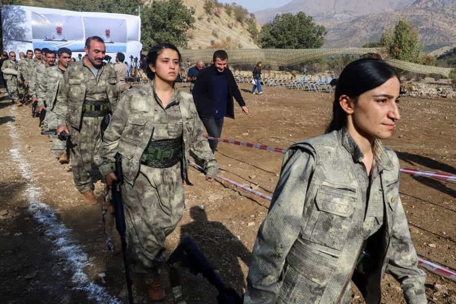 زنان و مردان عضو حزب کارگران کردستان (پ‌ک‌ک) در یک صف در حال حرکت‌اند. در کنار آن‌ها صندلی‌های زیادی دیده می‌شود.